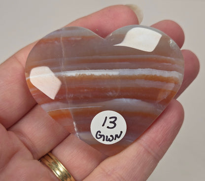 A heart-shaped polished agate stone with orange, brown, and white banded stripes, held in a person's hand. A small white sticker with the handwritten text '13 Green' is attached to the stone.