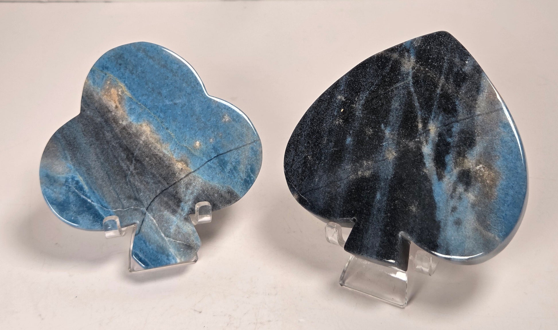 Two blue-spaced ace-of-spades playing card pendants made of polished gemstone, displayed on clear acrylic stands against a light marble background.