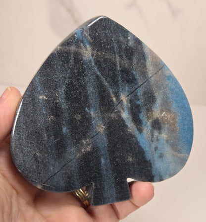 A hand holding a teardrop-shaped black and blue speckled stone object, likely a decorative piece or compact mirror, against a light marble-patterned background.
