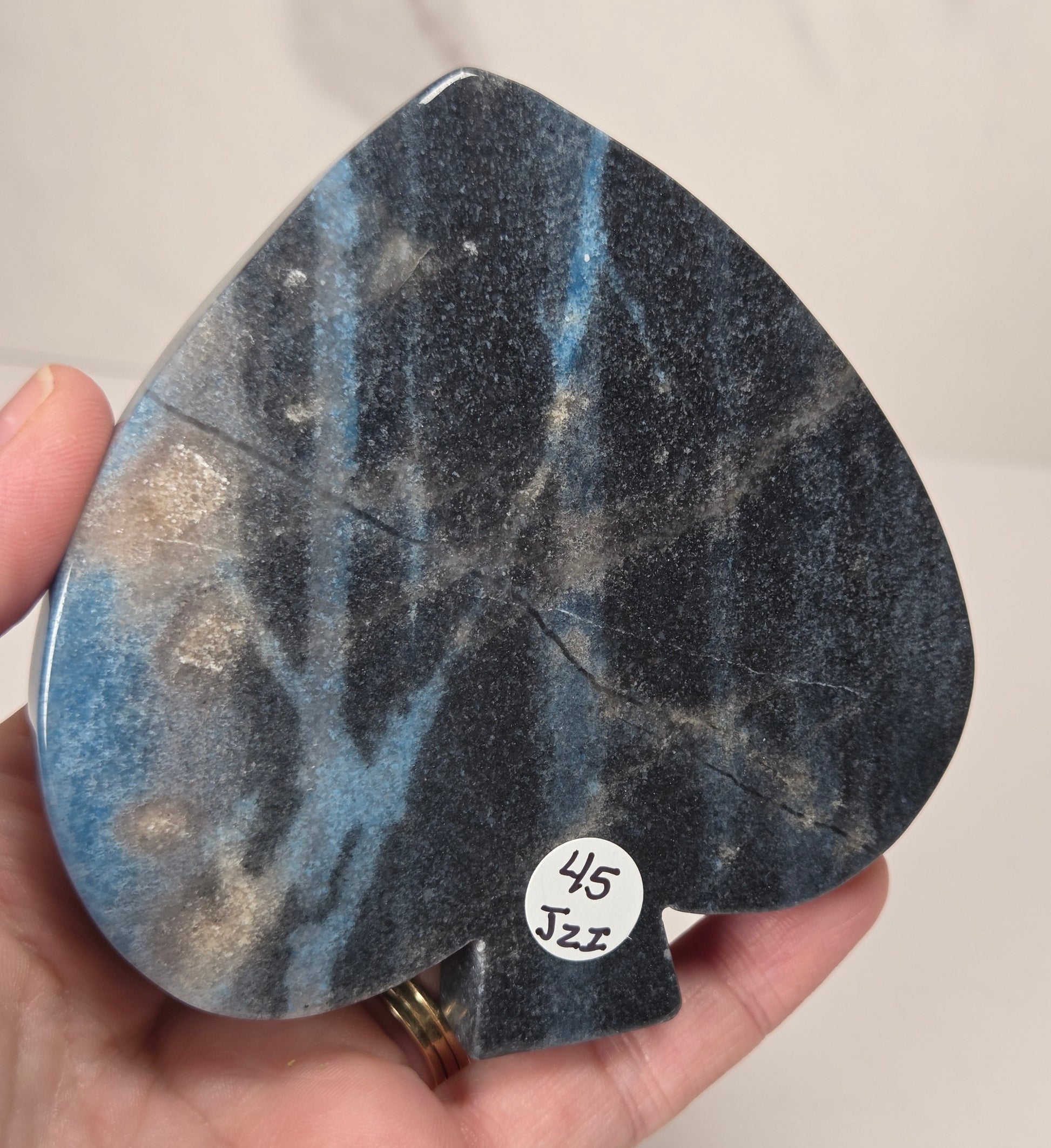 A hand holding a teardrop-shaped black and blue speckled stone object with a small white sticker reading '45 Jzz' on a light background.