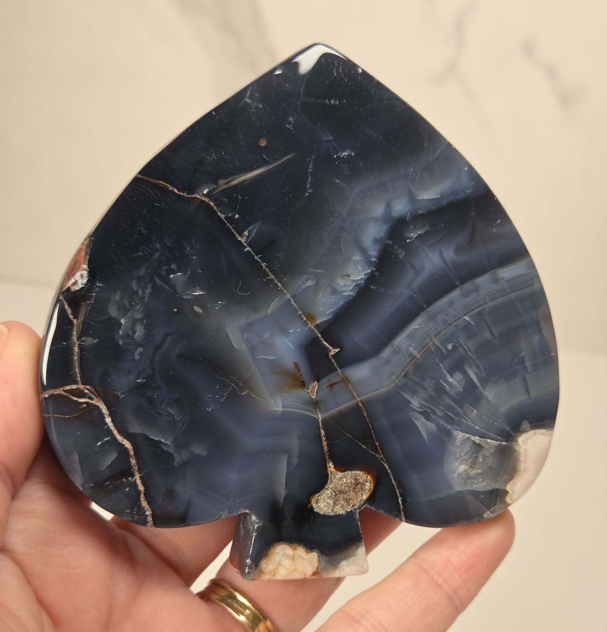 A hand holding a teardrop-shaped black agate stone with natural beige and white veining, set against a light marble background.