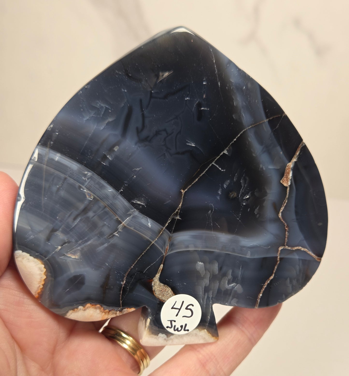 A hand holds a polished teardrop-shaped agate stone with dark blue, gray, and beige banding and natural fissures. A small white sticker with the handwritten text '45 Joul' is attached near the bottom.