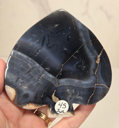 A hand holds a polished teardrop-shaped agate stone with dark blue, gray, and beige banding and natural fissures. A small white sticker with the handwritten text '45 Joul' is attached near the bottom.