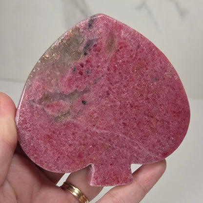 A hand holding a polished pink spade-shaped stone or crystal with natural marbling and inclusions, set against a light marble-patterned background.
