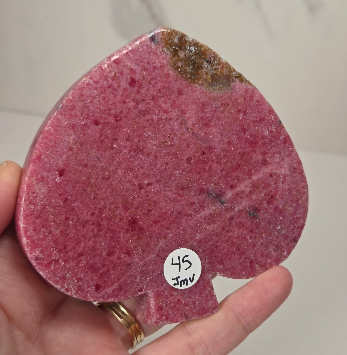 A hand holding a polished pink spade-shaped stone with a rough brown section near the top and a small white sticker reading '45 JMV' on the blade, against a light marble-patterned background.