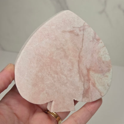 A hand holding a pinkish-white speckled stone playing card spade, likely rose quartz, with a gold ring on the finger, set against a light marble-patterned background.