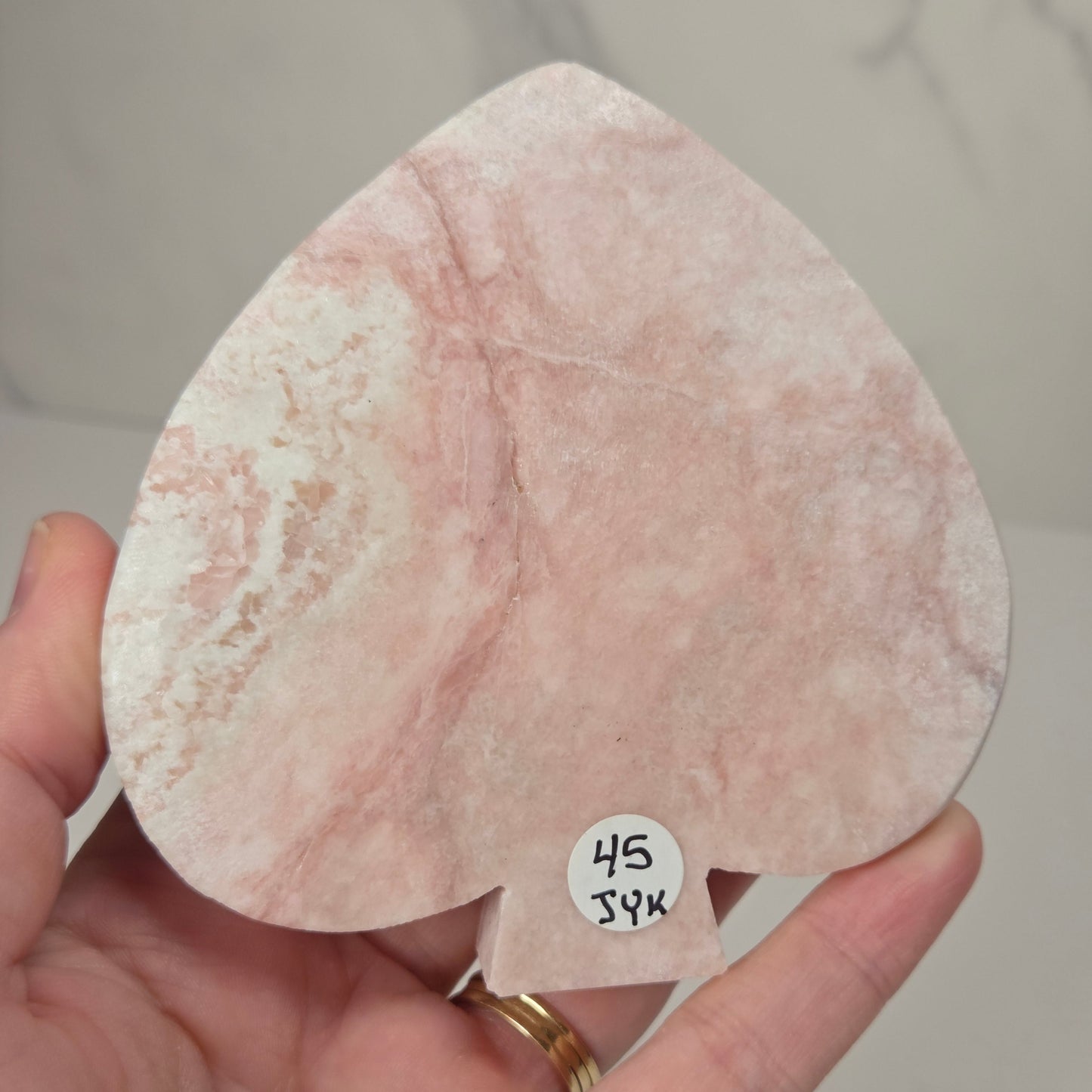 A hand holds a pink and white marble heart-shaped object with a small white price tag attached. The background is a light gray marble-patterned surface.
