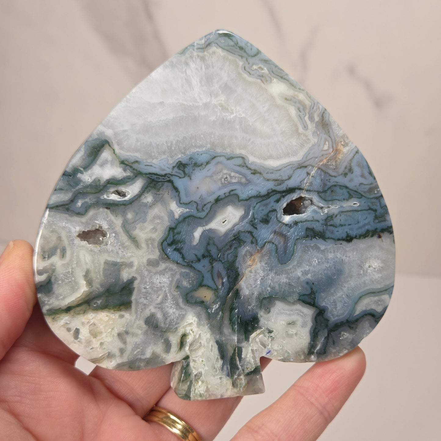 A teardrop-shaped agate stone cutout with a spade-like silhouette, held in a hand. The stone features natural marbled patterns of slate blue, forest green, white, and gray.