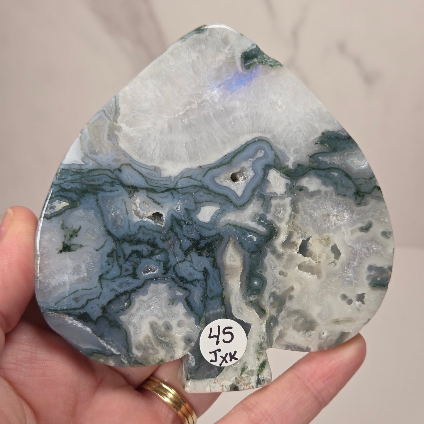 A hand holding a teardrop-shaped polished stone with swirling blue, gray, white, and green marbled patterns. A small white sticker with handwritten text is attached near the bottom.