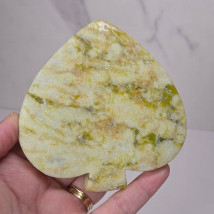 A hand holding a teardrop-shaped pipsqueck crystal gemstone with natural green and yellow marbling, set against a light gray marble background.