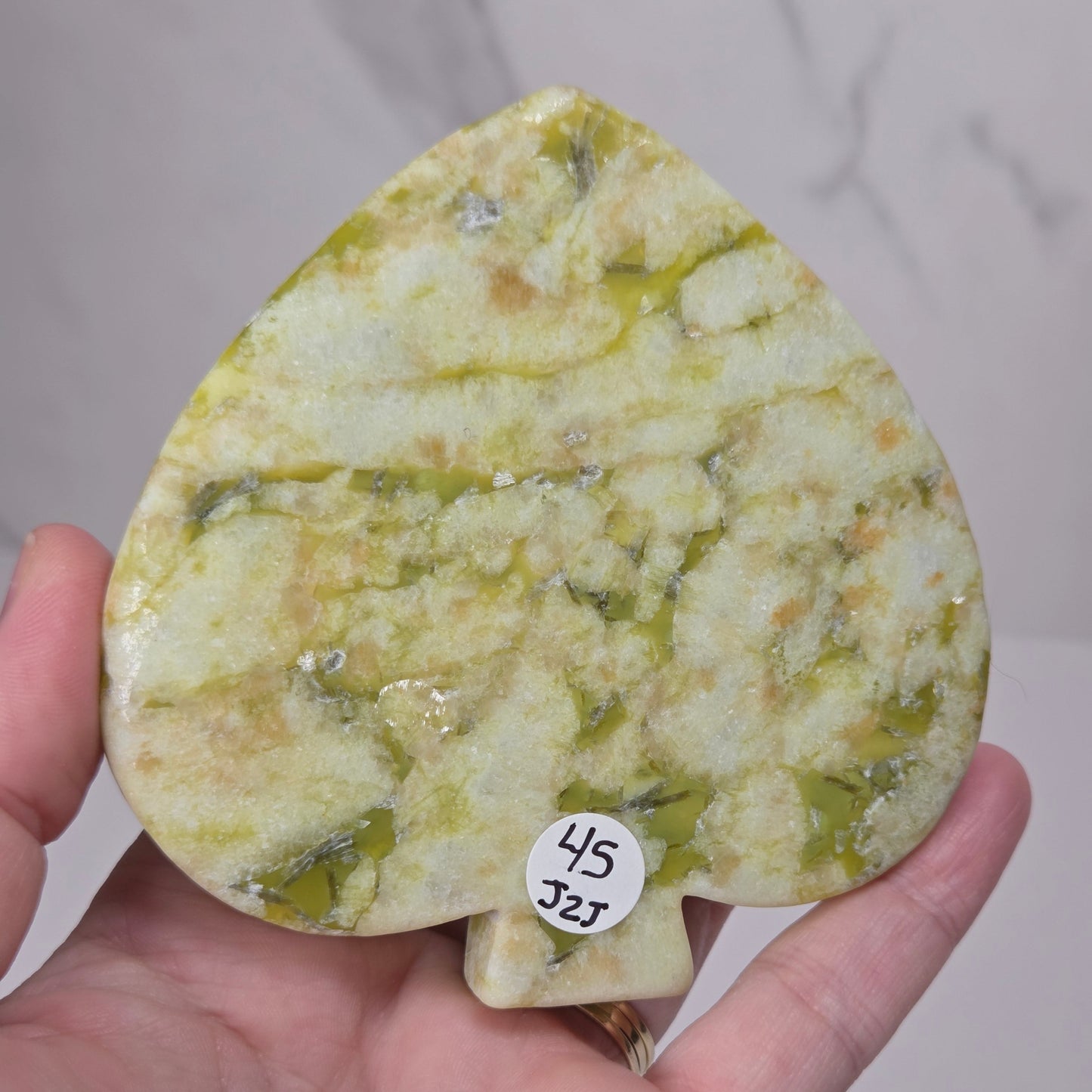 A hand holding a teardrop-shaped polished stone object with a marbled yellow-green and cream pattern, featuring a small white sticker with the numbers 45 and J25.