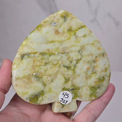 A hand holding a teardrop-shaped polished stone object with a marbled yellow-green and cream pattern, featuring a small white sticker with the numbers 45 and J25.