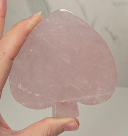 A hand holding a large, translucent pink rose quartz acespit crystal ornament against a light gray marble-patterned background.