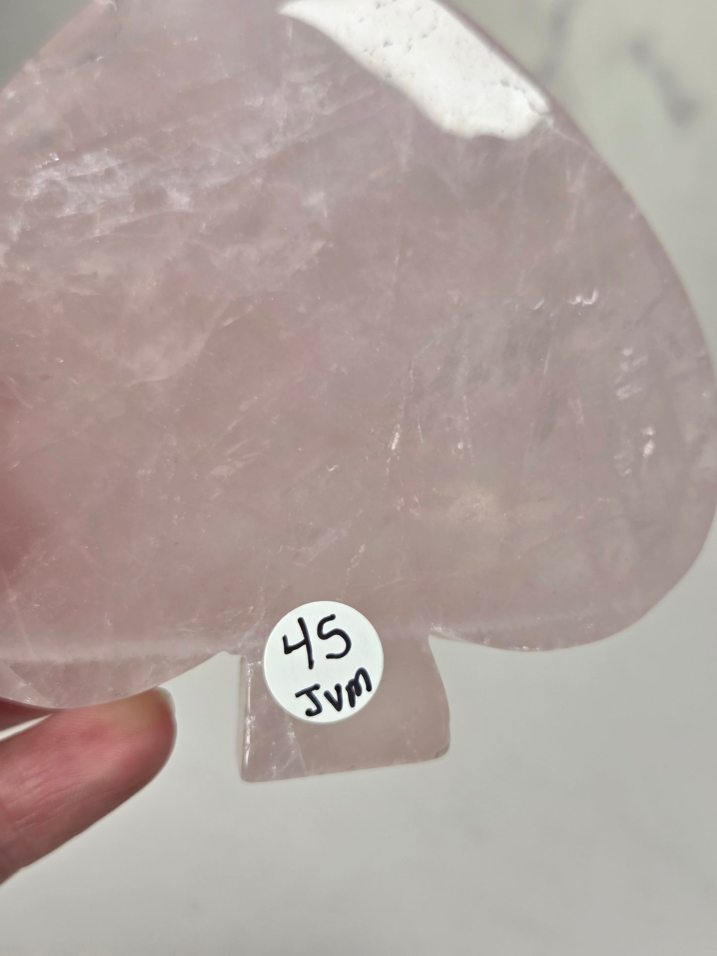A translucent pale pink teardrop-shaped crystal or stone piece held in a hand, with a small white sticker reading '45 JVN' attached near the bottom.