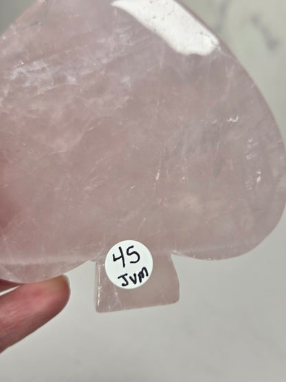 A translucent pale pink teardrop-shaped crystal or stone piece held in a hand, with a small white sticker reading '45 JVN' attached near the bottom.