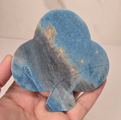 A hand holding a teardrop-shaped blue stone ornament with natural marbled gray and gold veining, set against a light beige marble background.