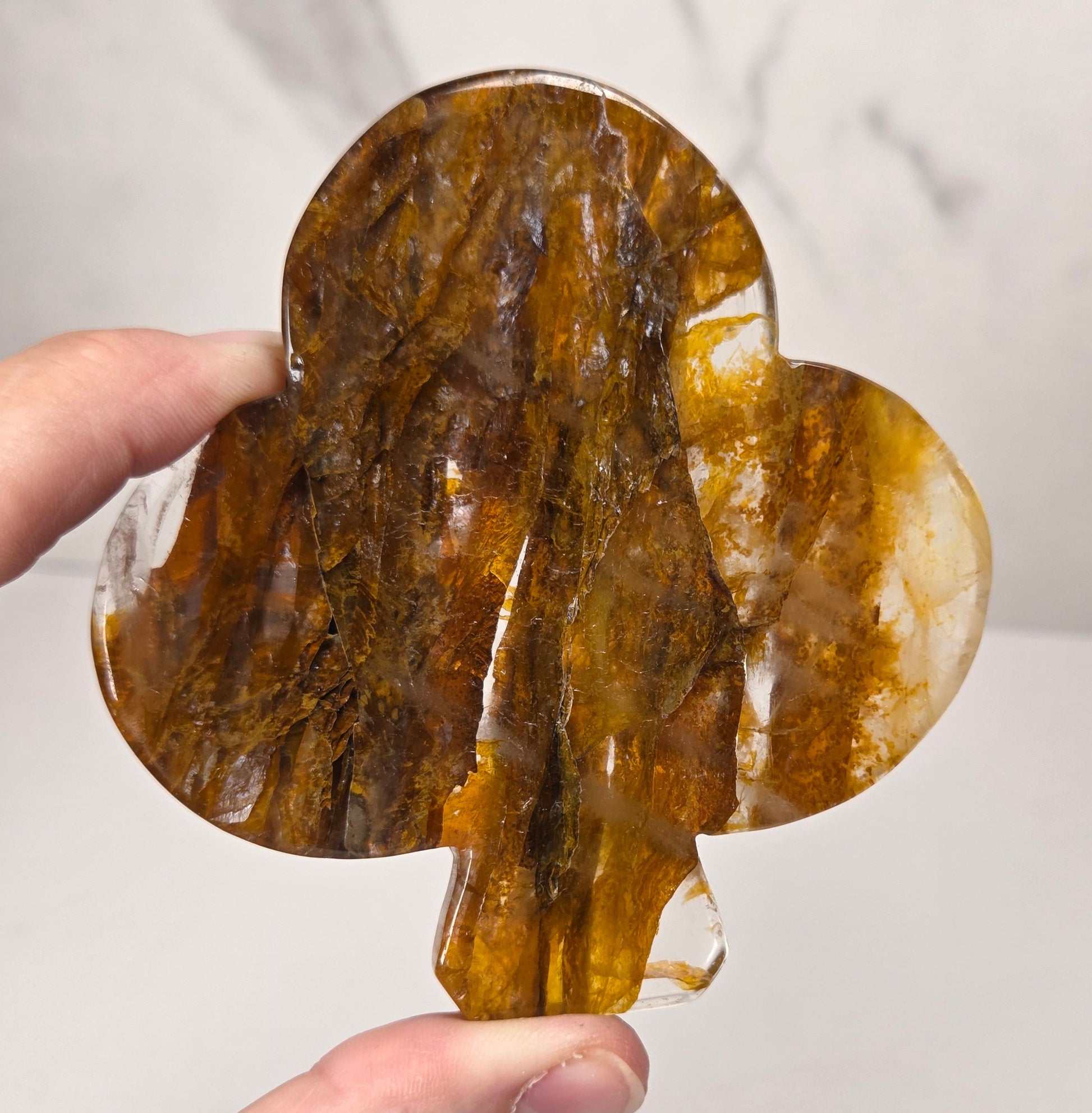 A hand holding a teardrop-shaped carved crystal or stone piece with a brown and amber marbled pattern, set against a light gray marble-patterned background.