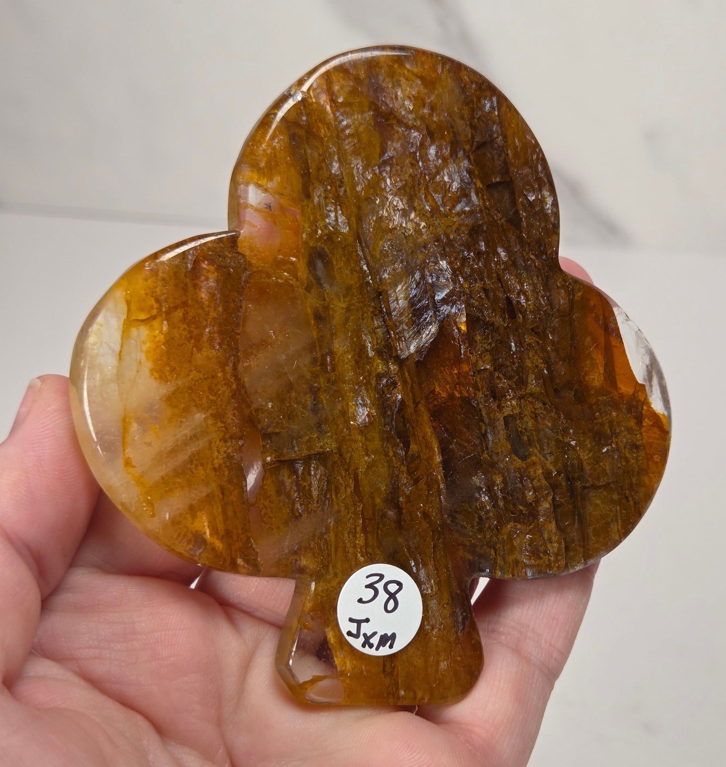 A hand holding a polished, heart-shaped brown gemstone with natural inclusions and a small white sticker reading '38 5cm'.