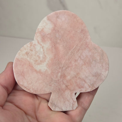 A hand holding a pink stone playing card suit charm shaped like a club, featuring natural pink and white marbled patterns.