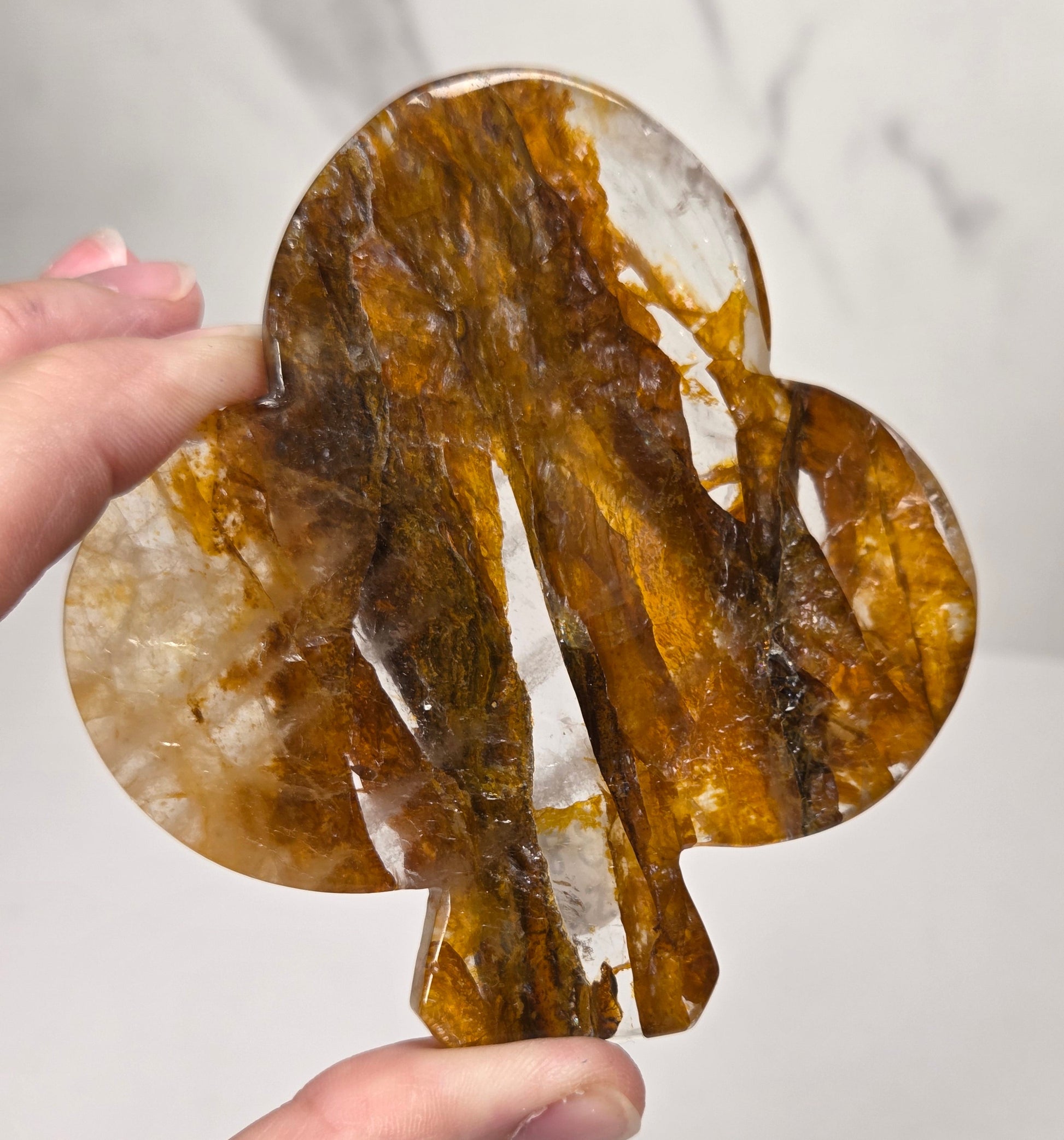 A hand holding a polished, tree-shaped crystal or stone with rich amber-brown and translucent white marbling, set against a light gray marble-patterned background.