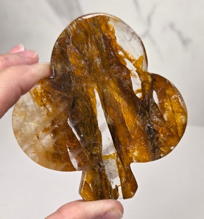 A hand holding a polished, tree-shaped crystal or stone with rich amber-brown and translucent white marbling, set against a light gray marble-patterned background.