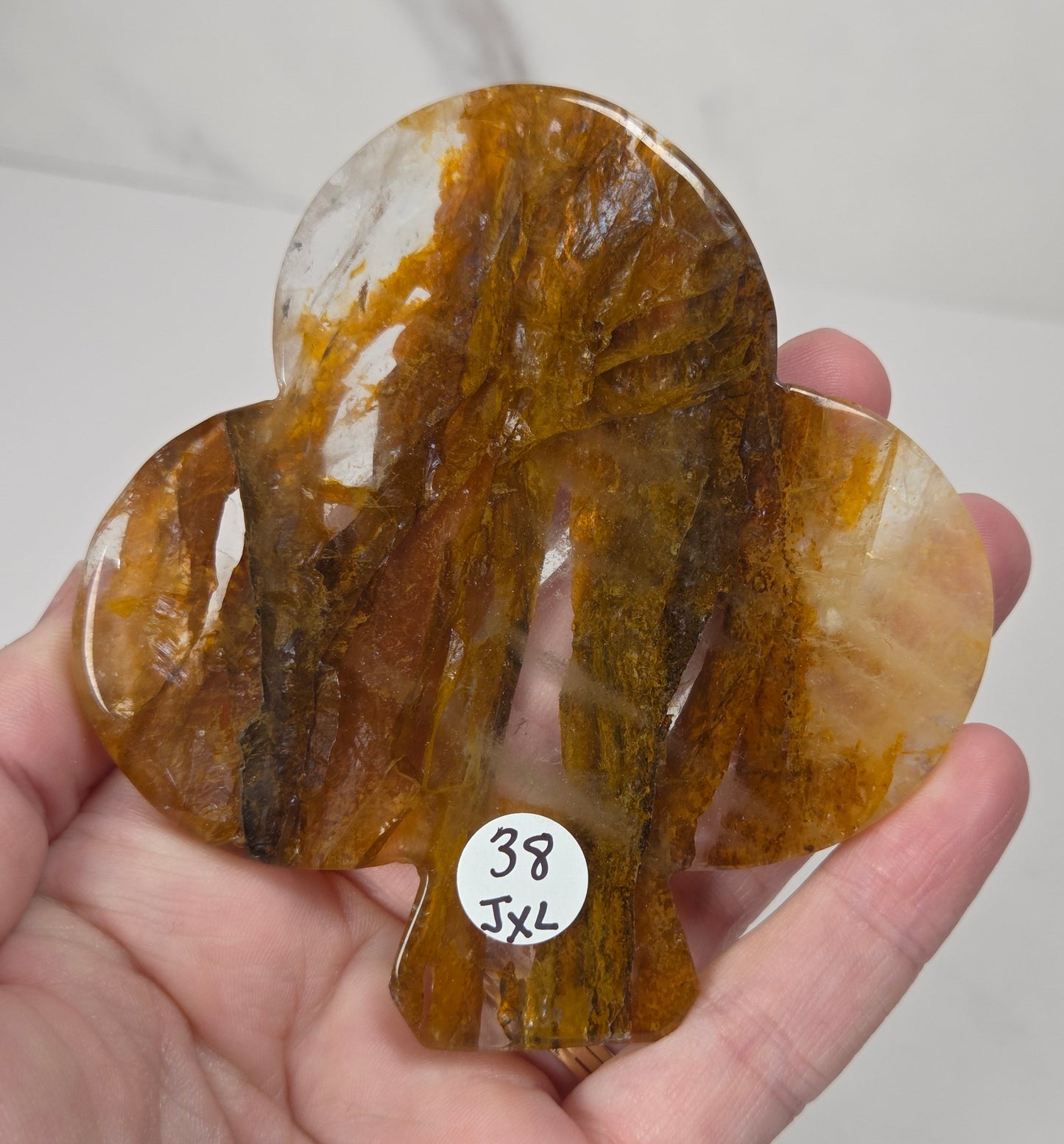 A hand holds a carved organic-shaped translucent stone piece with swirling amber, honey, and brown tones over a light marble-like background. A small white sticker reads '38 JXL'.
