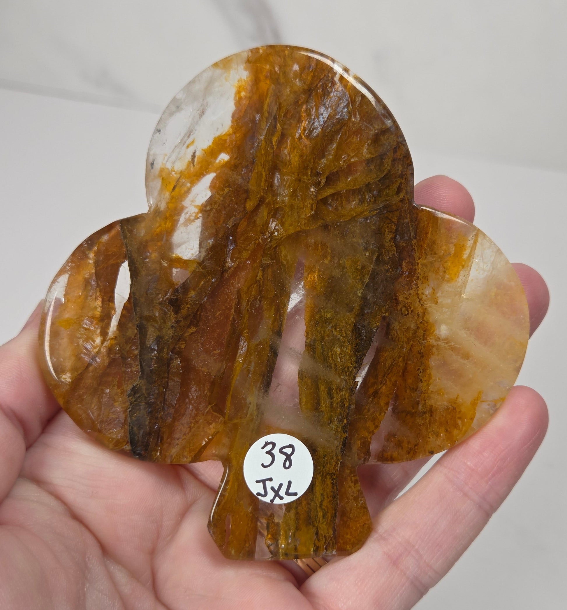 A hand holds a carved organic-shaped translucent stone piece with swirling amber, honey, and brown tones over a light marble-like background. A small white sticker reads '38 JXL'.