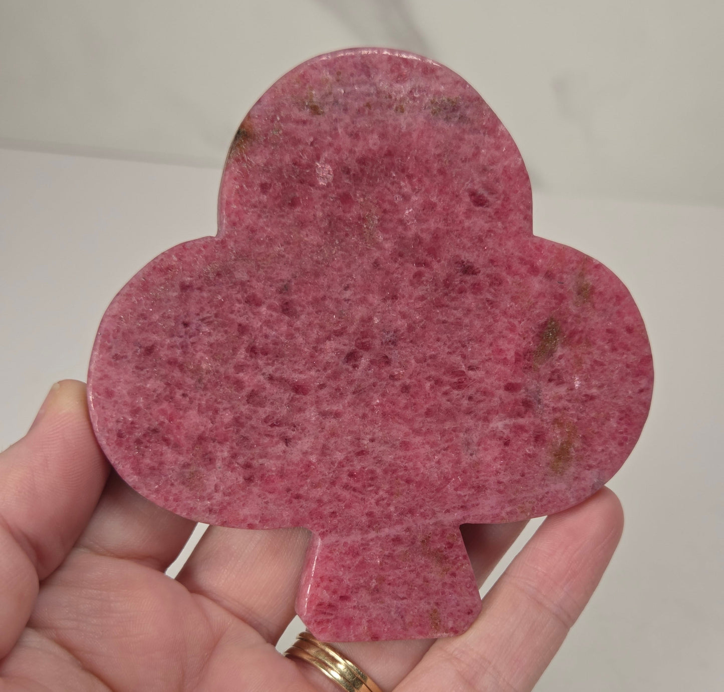 A hand holding a flat, club-shaped pink stone or crystal with a speckled texture and small brown inclusions, against a light neutral background.