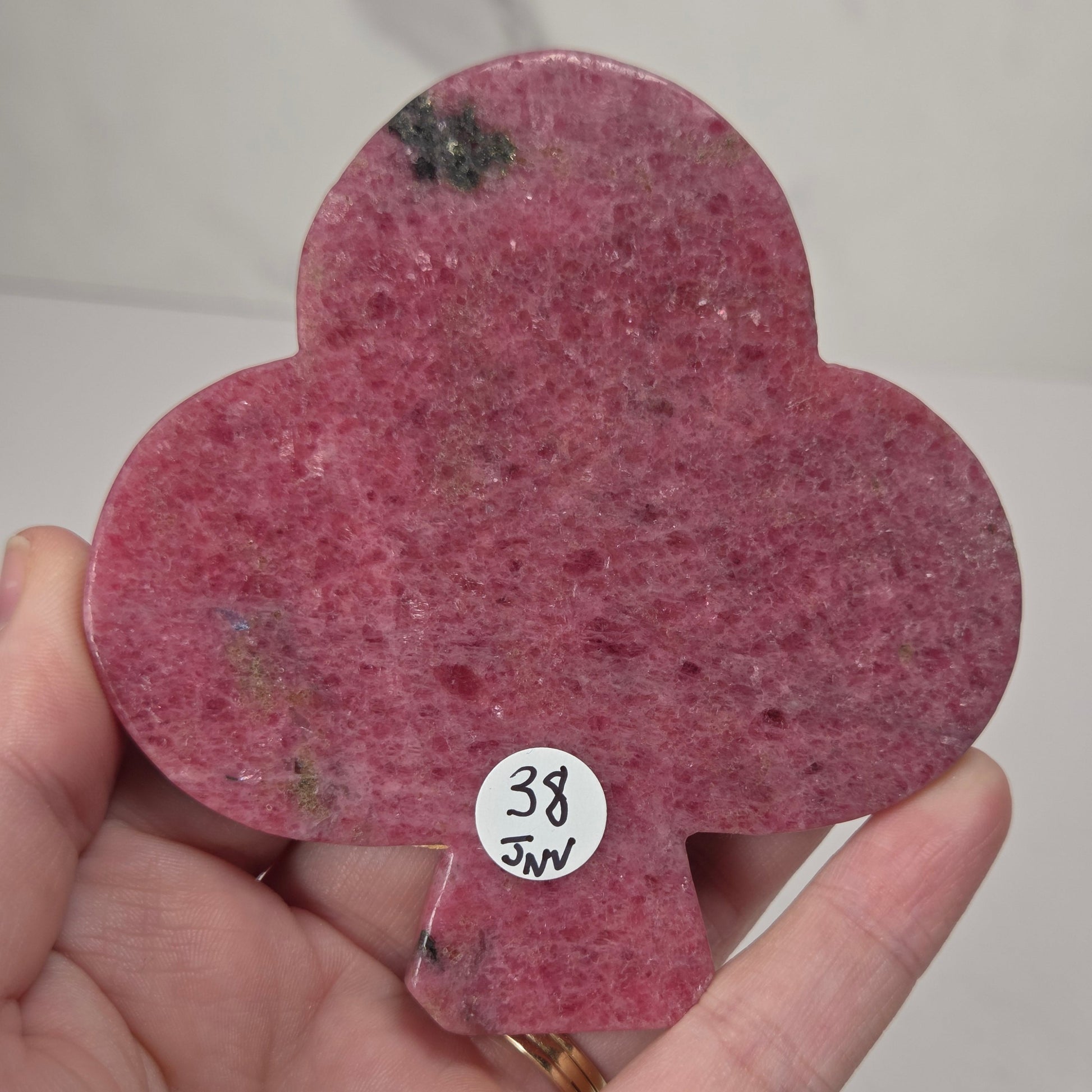 A hand holding a teardrop-shaped pink stone specimen with a small white sticker reading '38 JNW' on it, against a light gray background.