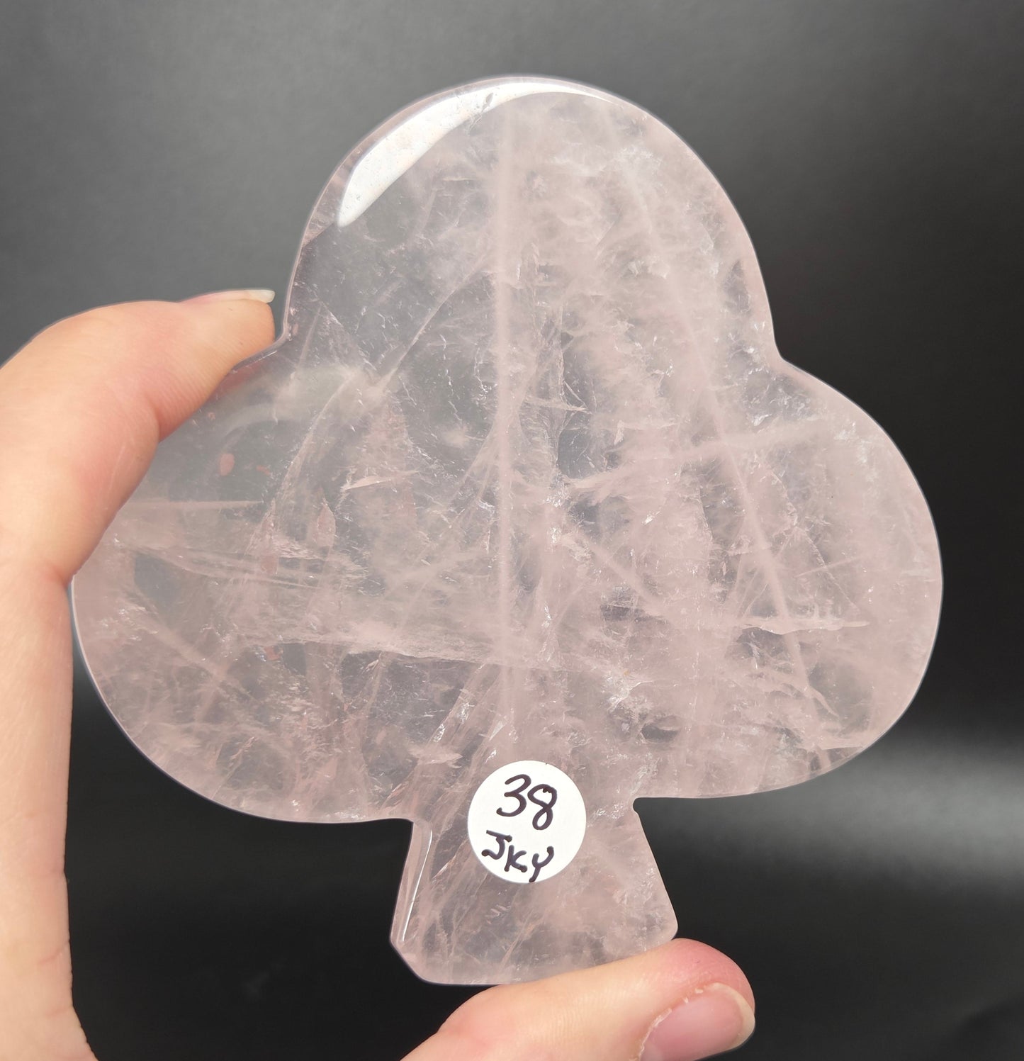 A hand holding a translucent pale pink teardrop-shaped crystal pendant with dense fibrous inclusions, photographed against a dark gray background. A small white sticker on the crystal reads '38 Jkv'.