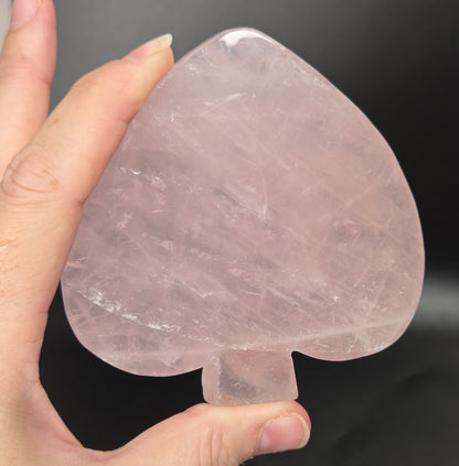 A hand holding a translucent pink rose quartz playing card spade-shaped crystal pendant against a dark gray background.