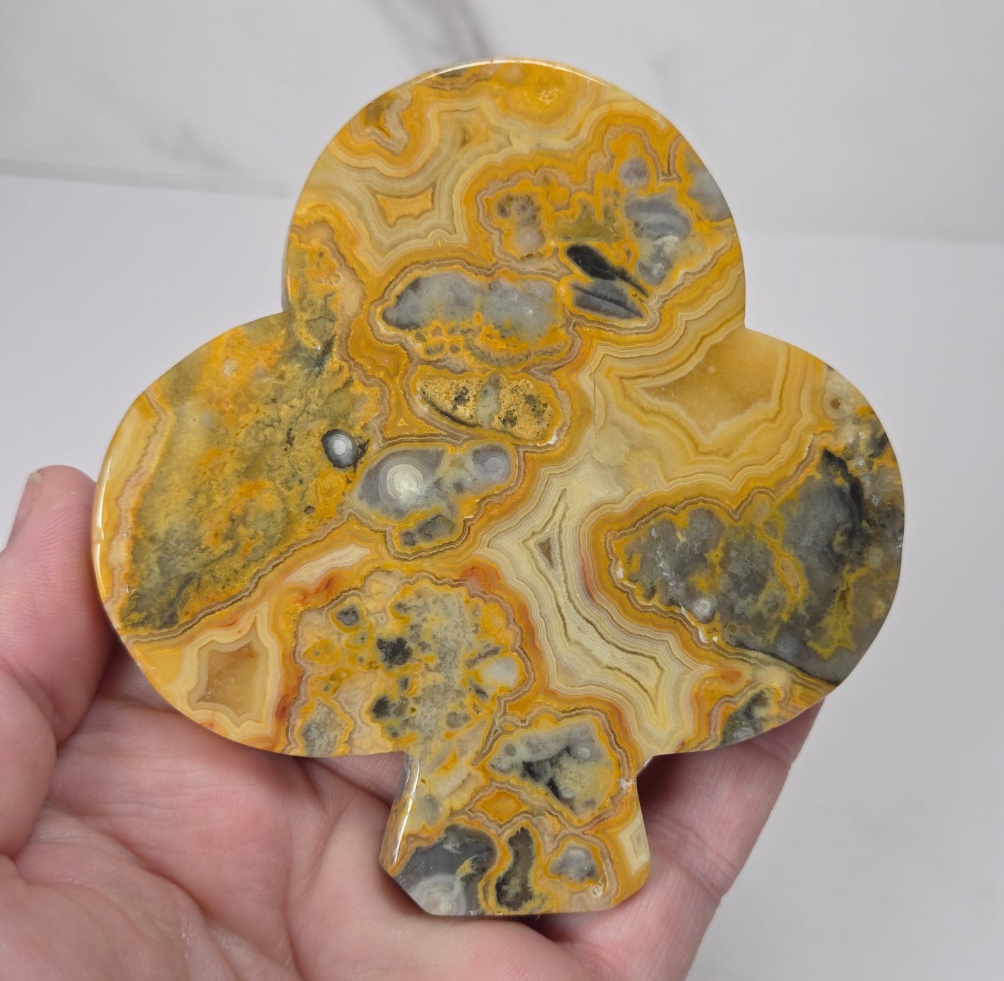 A hand holding a three-lobed decorative object made from polished agate stone with swirling bands of yellow, orange, gray, and cream. The object is shown against a light gray marble-patterned background.
