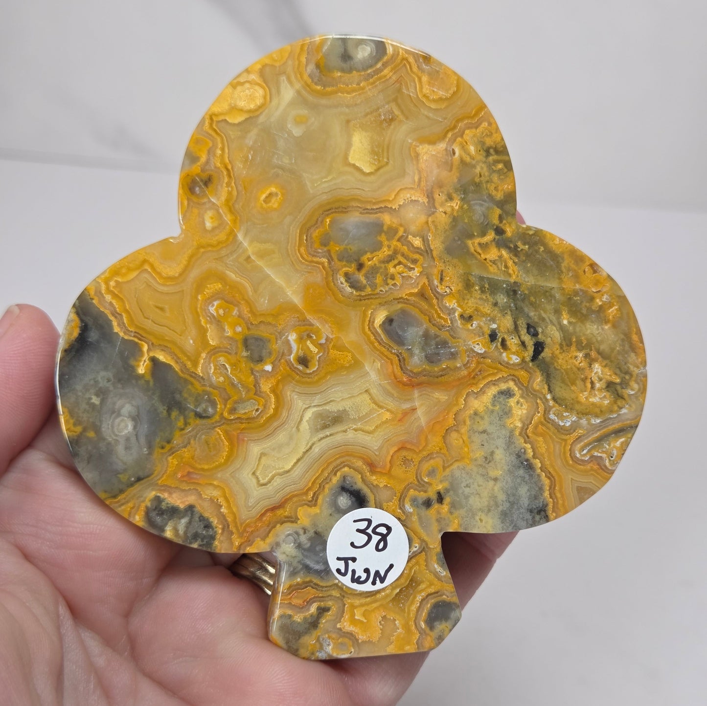 A hand holding a teardrop-shaped piece of yellow and gray agate stone with swirling band patterns, featuring a small white sticker that reads '38 Jun'.