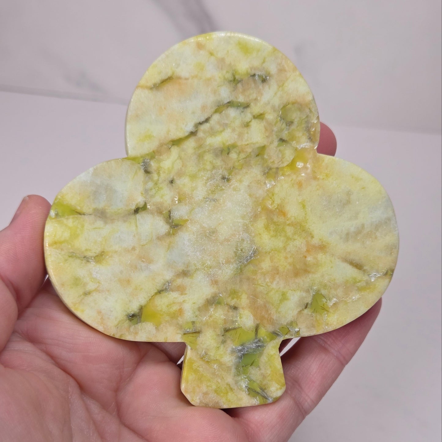 A hand holding a polished yellow-green gemstone cut in the shape of a club, showing natural marbling and inclusions.