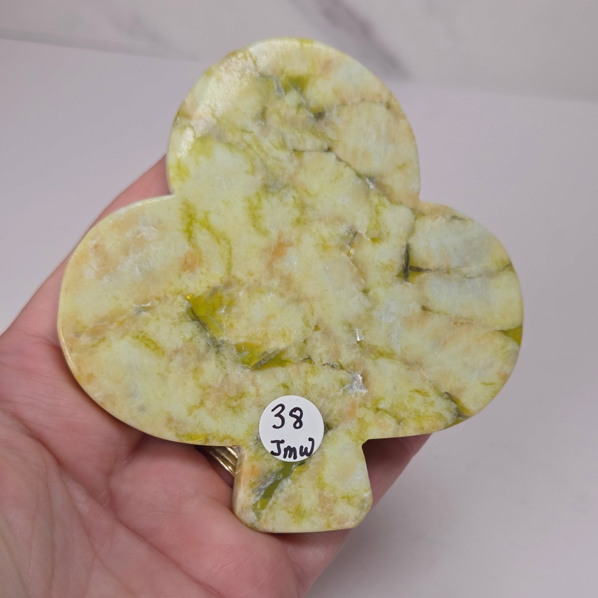 A hand holding a small, polished, clover-shaped stone with a marbled yellow and green pattern. A small white sticker with the handwritten text '38 Jmw' is attached to the bottom of the stone.