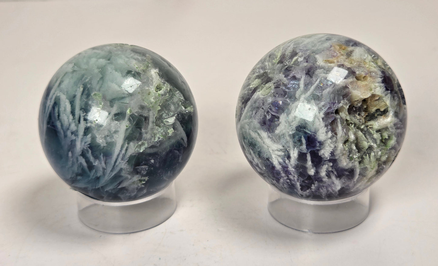 Two polished crystal spheres on clear display stands, one with blue-green and white marbling and the other with purple, green, and gold veining, set against a light marble-patterned background.