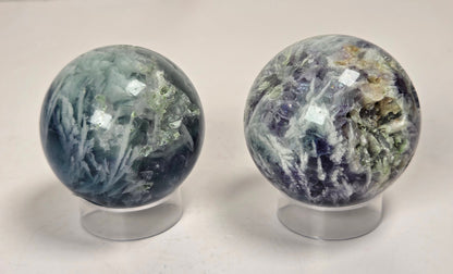 Two polished crystal spheres on clear display stands, one with blue-green and white marbling and the other with purple, green, and gold veining, set against a light marble-patterned background.
