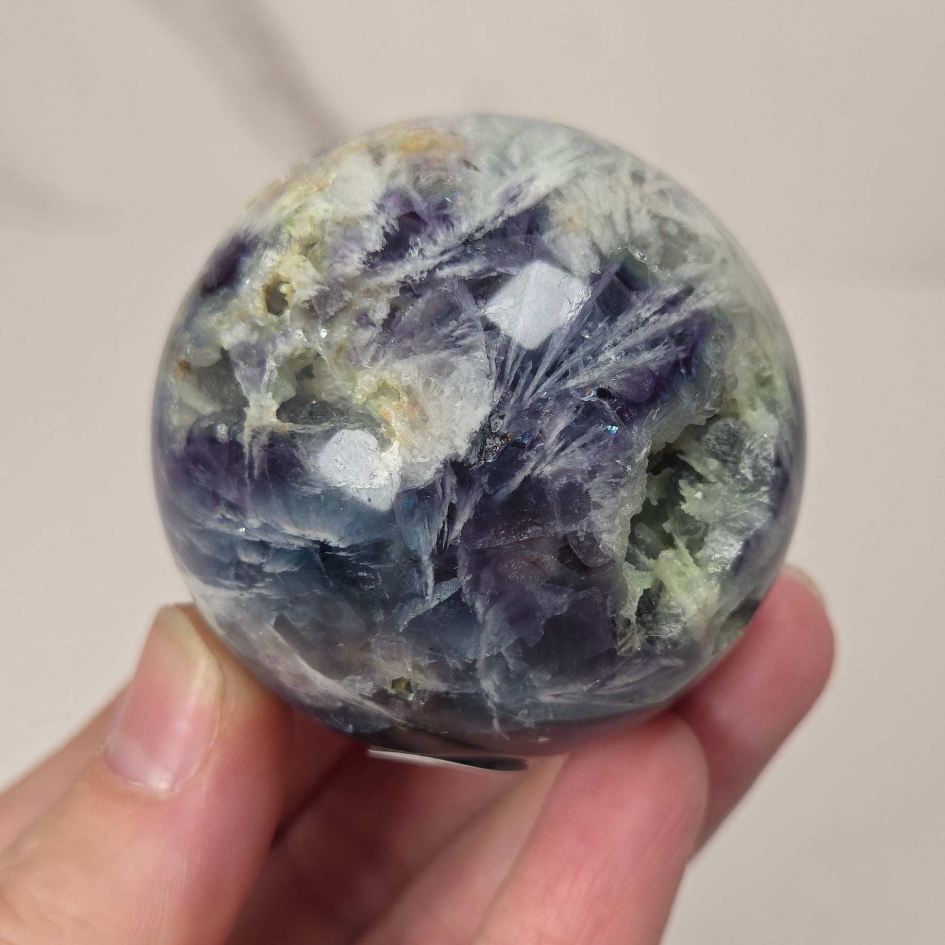 A hand holding a polished round gemstone with natural crystalline patterns in shades of blue, purple, gray, and white.