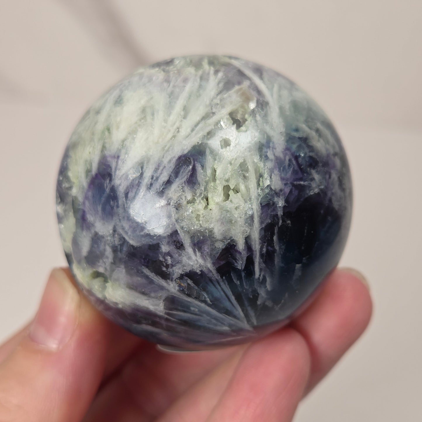 A hand holding a polished round crystal ball with swirling patterns of dark blue, purple, white, and light green.