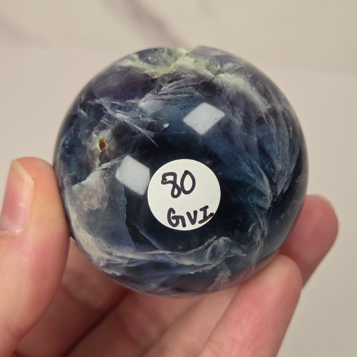 A hand holding a polished round gemstone with a marbled dark blue, gray, and white pattern. A small white sticker with handwritten text is attached to the front of the stone.