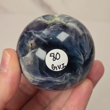 A hand holding a polished round gemstone with a marbled dark blue, gray, and white pattern. A small white sticker with handwritten text is attached to the front of the stone.