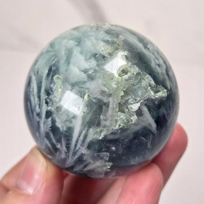 A hand holding a polished round crystal sphere with white and light green mineral inclusions against a light marble-like background.