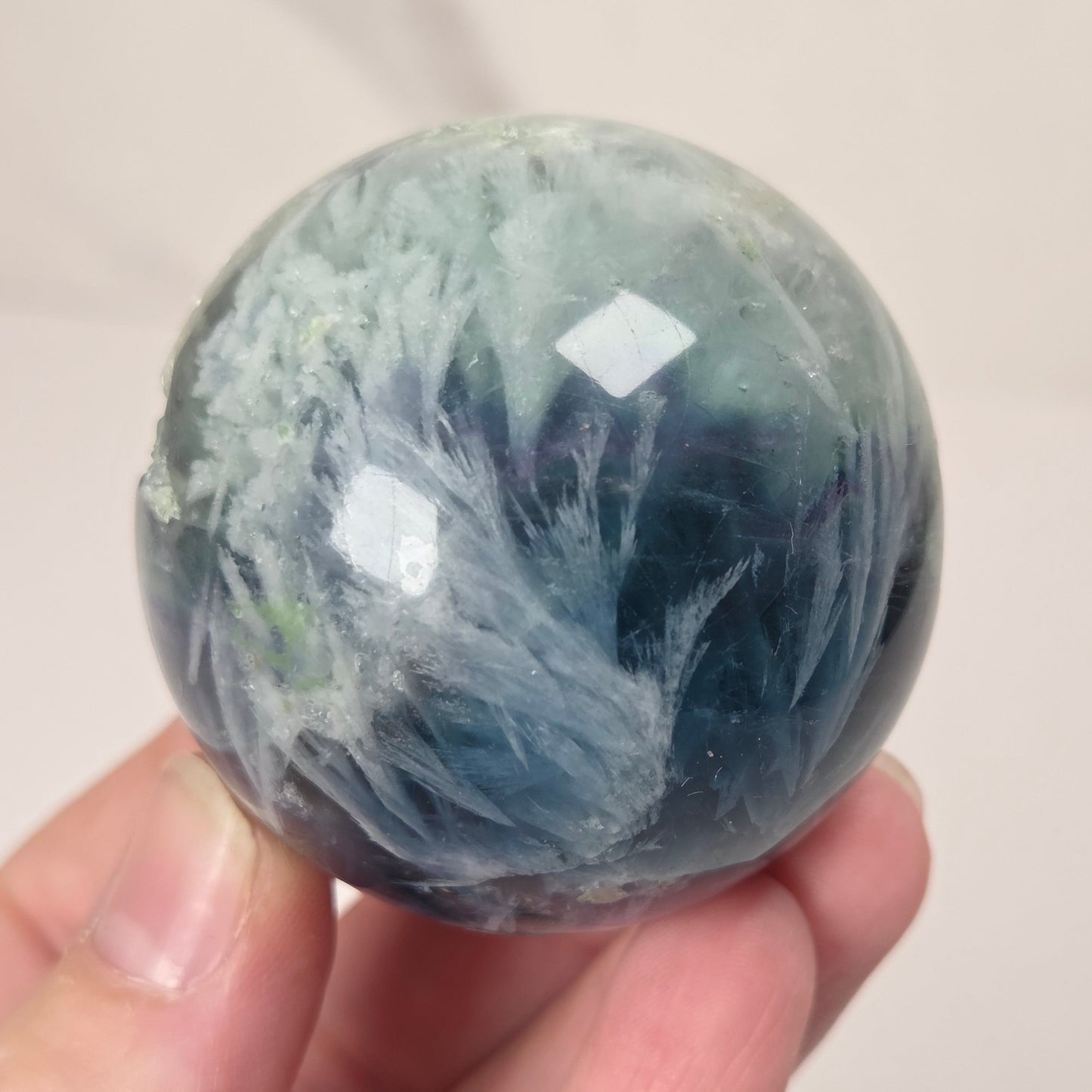 A hand holding a polished round crystal sphere with a marbled pattern of blue, green, and white.