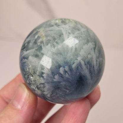 A hand holding a polished round crystal sphere with blue-gray and white mineral or crystal formations, against a soft light beige background.