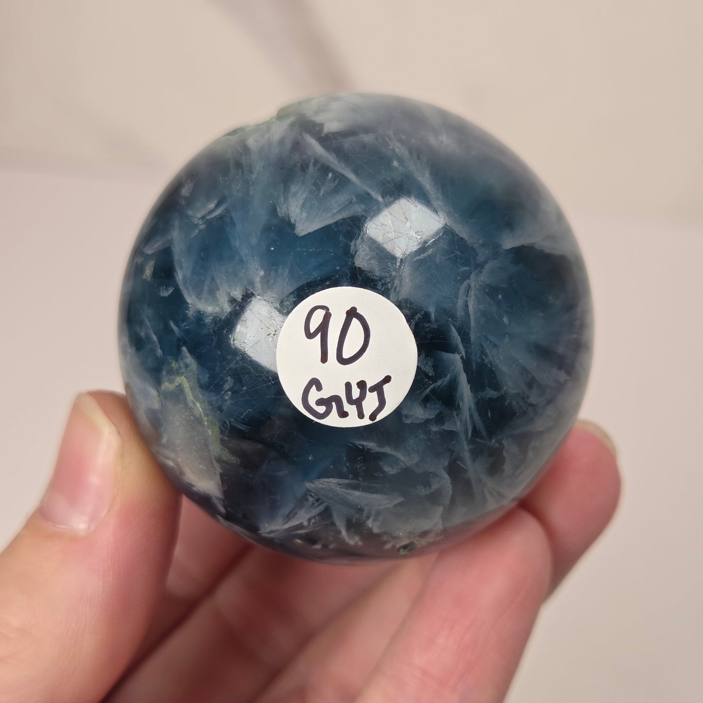 A hand holding a polished blue fluorite sphere with a white circular sticker that reads '90 GHS'.