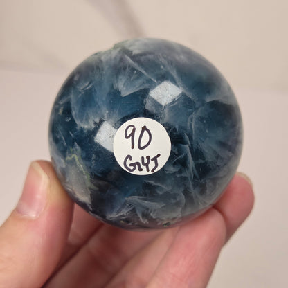A hand holding a polished blue fluorite sphere with a white circular sticker that reads '90 GHS'.