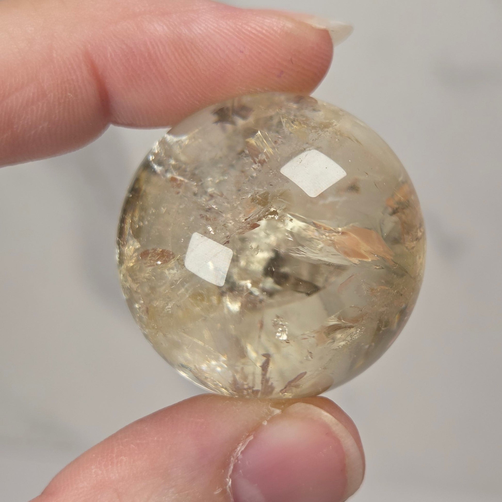 A hand holding a faceted, round clear gemstone with internal inclusions against a light gray background.