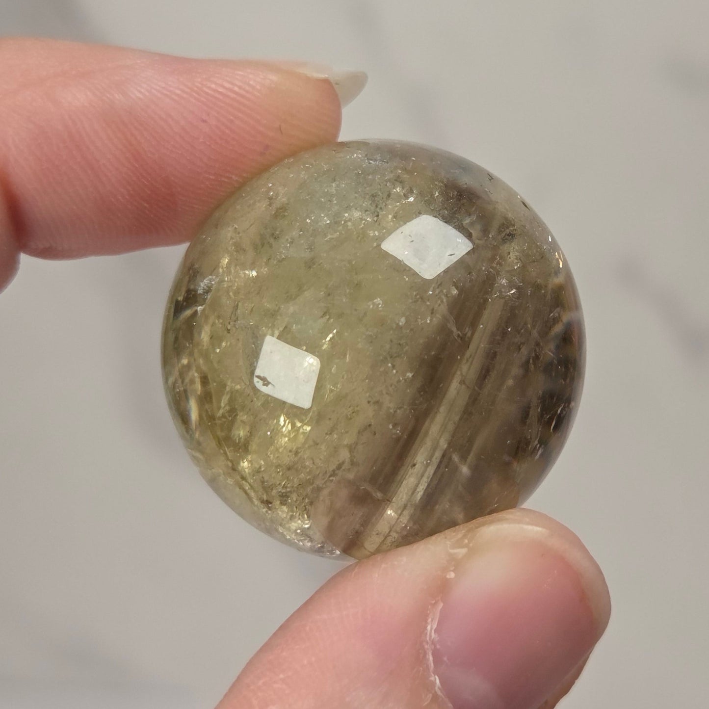 A hand holding a small, round, faceted clear stone bead or cabochon with internal inclusions, set against a light gray marble-patterned background.
