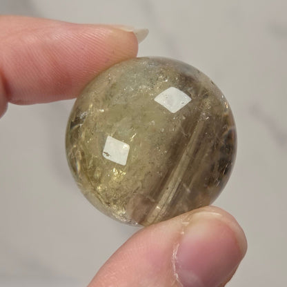 A hand holding a small, round, faceted clear stone bead or cabochon with internal inclusions, set against a light gray marble-patterned background.