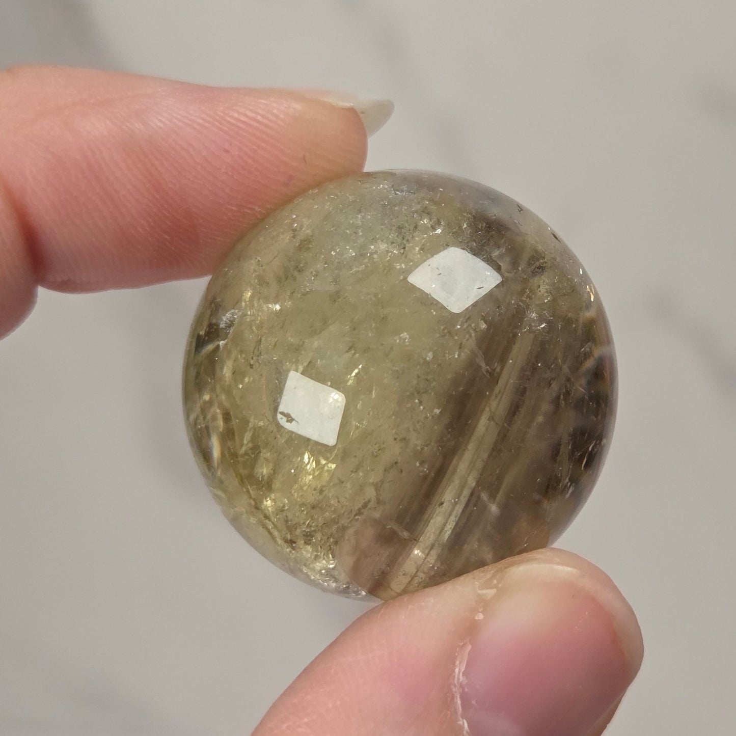 A hand holding a small, round, faceted clear stone bead or cabochon with internal inclusions, set against a light gray marble-patterned background.